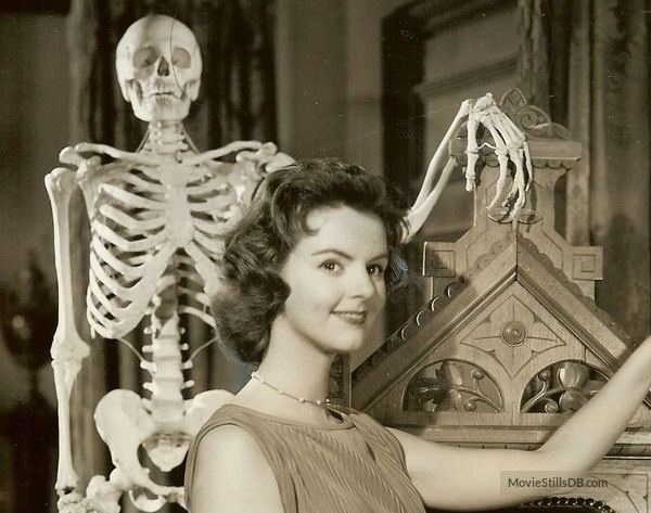 Carolyn Craig smiling beside a human skeleton, with short wavy hair, wearing a necklace, and a sleeveless top.