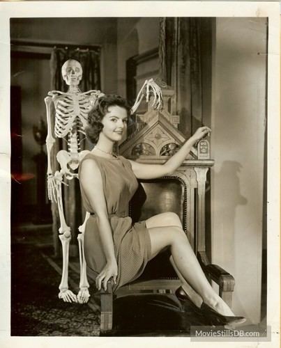 Carolyn Craig smiling while sitting on a chair beside a human skeleton, with short wavy hair, wearing a sleeveless dress, and black shoes.