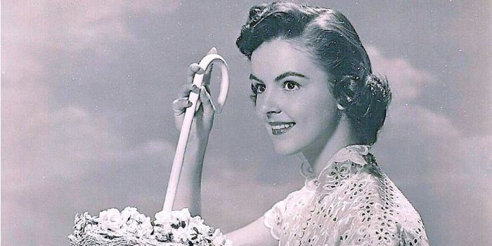Carolyn Craig with a bright smile while holding a cane's handle with flowers, with short hair, and wearing a white lace blouse.