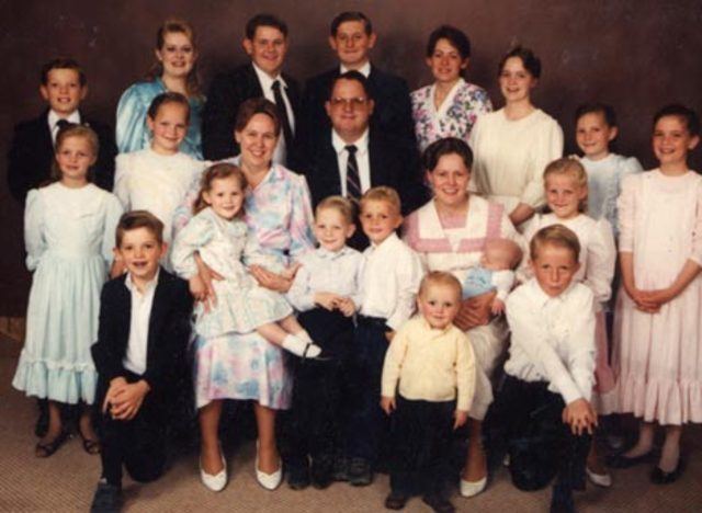 A boy on left (first row) is smiling, has blonde hair, both hands on his knee, and wears white long sleeves under a black suit, and black pants. 2nd from left is a woman smiling, sitting down while carrying a baby girl in her right hand, has blonde hair, and wears a colored pink-blue dress and white shoes, a baby girl on her lap is smiling, has brown hair, wears light blue long sleeves under a white dress, white socks and black shoes. 3rd from left is a boy, smiling, has blonde hair, both hands holding each other, and wears white long sleeves, black pants, and black shoes. 4th from left is a boy smiling, has blonde hair, left hand inside his left pocket, wears white long sleeves, black pants with pocket, and black shoes. 5th from left is a woman smiling, has black hair, sitting down while carrying a baby in her right hand, wearing a pink-white dress and white shoes,  a baby in her right hand is sleeping, bald, and wearing a white-blue dress. In front is a boy smiling, bald, wearing yellow long sleeves, black pants, and black shoes, beside him is a boy, serious, has blonde hair, left knee is bent, both hands on his left knee, wears white long sleeves, black pants, and black shoes. 6th from left is a girl smiling, has blonde hair, and wears a white long-sleeve dress, and white shoes. On right is a girl smiling, both hands holding each other, wearing a pink long-sleeve dress. A girl on left (second row) is smiling, has brown hair, both hands on her back, and wears a blue long-sleeve dress, and black shoes. 2nd from left is a girl smiling, has blonde hair, and wears a white long-sleeve dress. In the middle is a man smiling, has black hair, wearing brown sunglasses, white long sleeves, and a black necktie with lines under a black suit. 3rd from left is Carolyn standing, has black hair, both hands holding each other, and wearing a white long-sleeve dress. On right is a girl smiling, has black hair, wearing a blue long sleeve dress. A boy on left (third row) is smiling, has black hair, and wears white long sleeves, a black necktie under a black suit, pants, and black shoes. 2nd from left is a woman smiling, has blonde hair, and wears a silk blue dress. 3rd from left is a man smiling, has black hair, and wears white long sleeves, a black necktie under a black suit. 4th from left is a man smiling, wearing white long sleeves under a black suit. 5th from left is a woman smiling, has black hair, wearing a colored dress.