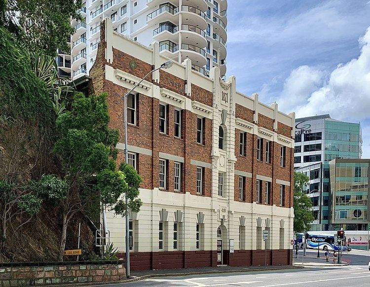 At the top is a blue sky and white clouds, at the left is a hillside with large roots, trees and bushes, in the middle is Castlemaine Perkins building has white wall roof, a brown brick wall, and a white painted wall with dark brown at the bottom in the ground floor in front is a highway and a white tall tall building at the back, at the right is a cyan building with large mirror windows and a blue bus.