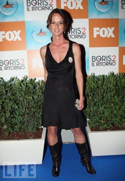 Caterina Guzzanti smiling, holding a pack of cigarettes, and standing in front of the Boris 2 IL Ritorno and Fox backdrop with tied-up hair and wearing black boots, earrings, and a black sleeveless dress with a low-cut neckline that exposes her cleavage