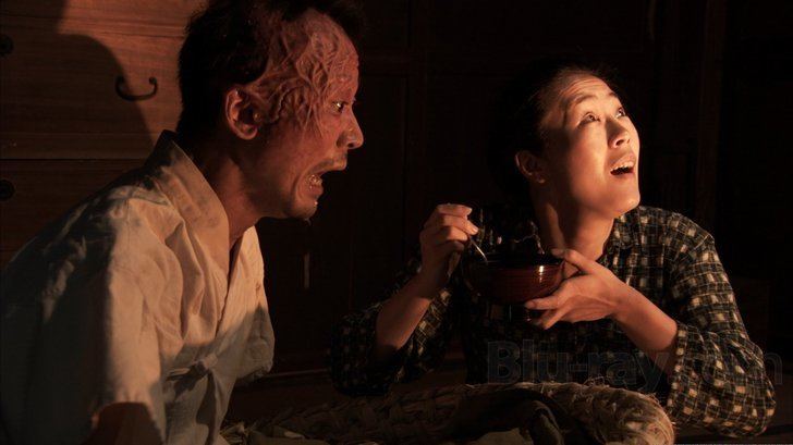 In the movie scene of Caterpillar 2010, from left is Keigo Kasuya, shocked with his mouth open while sitting on the bed, has burnt face from right ears to his forehead and has black hair wearing white shirt, at the right is Shinobu Terajima surprised, looking above at her right side, mouth half open sitting while feeding Keigo Kasuya with her right hand holding a spoon and left hand with a miso cup, has long black hair wearing a black and white checkered long sleeve clothes.
