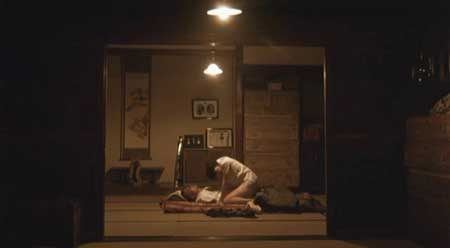 In the movie scene of Caterpillar 2010, Shinobu Terajima is on top of Keigo Kasuya with her both hand on floor kneeling has black hair wearing a white clothes on top, at the bottom is Keigo kasuya, lying on the bed looking at Shinobu Terajima above him, has burnt face black hair and no limbs wearing a white shirt.