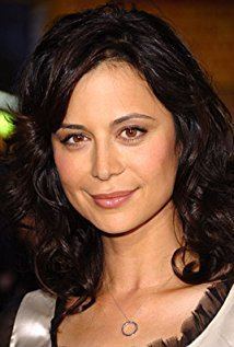 Catherine Bell with a tight-lipped smile and black curly hair while wearing a black and white blouse and necklace