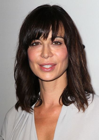 Catherine Bell smiling with a black wavy shoulder-length hair while wearing a white blouse