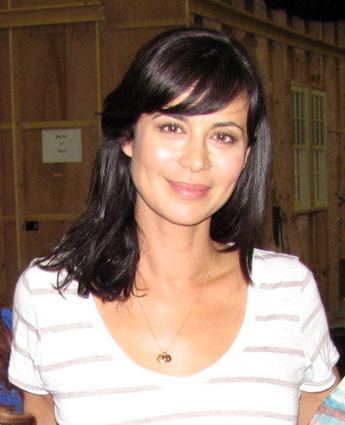 Catherine Bell with a tight-lipped smile and black shoulder-length hair while wearing a white and gray striped blouse and necklace