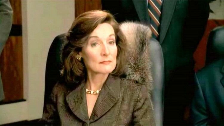 Catherine McGoohan looking at someone while sitting on a black office chair and wearing a brown coat, earrings, and necklace