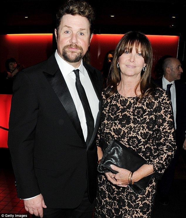 Cathy McGowan smiling and wearing earrings, a black floral dress, and carrying a black bag together with Michael Ball with a beard and mustache and wearing a black suit and a black tie.