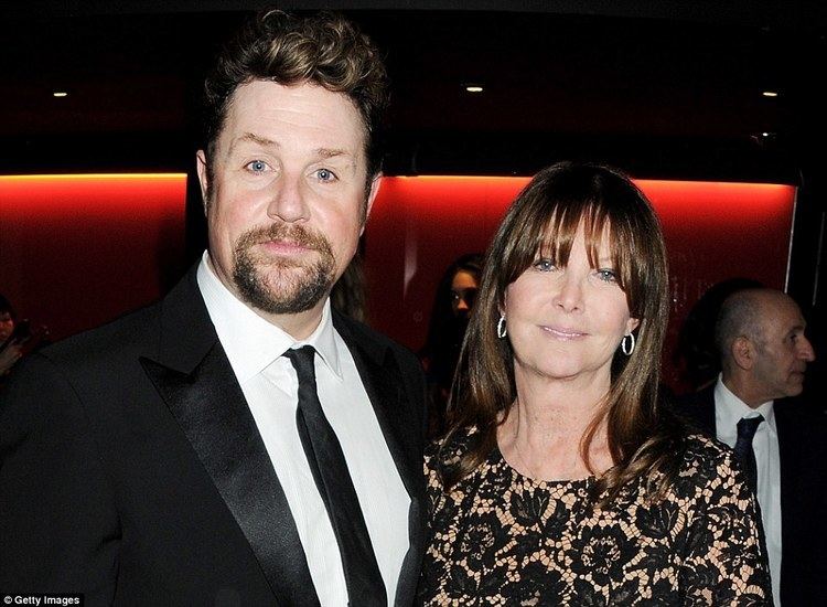 Cathy McGowan smiling and wearing earrings and a black floral dress together with Michael Ball with beard and mustache and wearing a black suit and a black tie.