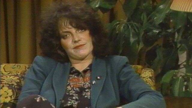 Cathy Smith sitting on a couch wearing a black printed shirt and a coat