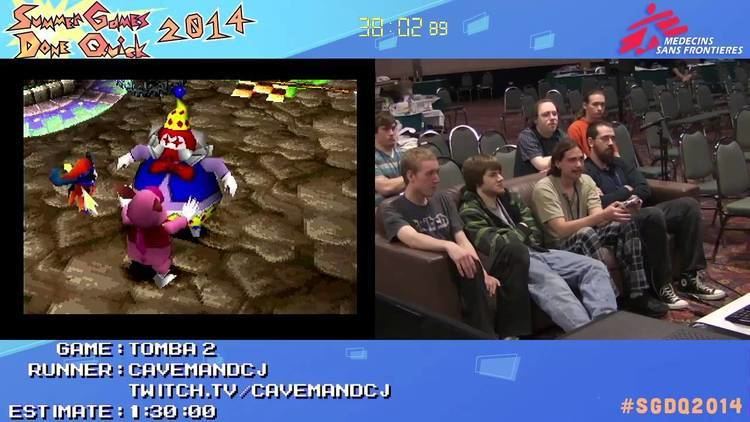 On the left, Tomba 2 game. On the right, Caveman DCJ sitting on a couch with Chibi and other players watching him playing Tomba 2. Caveman DCJ wearing a light green shirt and checkered black and white pants.