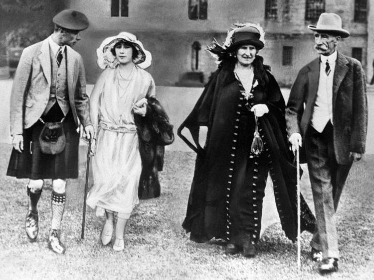 George VI on left is serious, has black hair, his left hand holding a cane, wearing a black hat, white long sleeves, a black necktie, a gray vest under a suit, a black skirt, a belt bag, and black shoes. 2nd from left is Lady Elizabeth Bowes-Lyon is serious, has black hair, left hand holding a black coat, wearing a white hat with a black design, necklace, a white dress, and white shoes. On right is Cecilia Bowes-Lyon, serious, left hand holding a small bag, wearing a black hat, black dress under a black and white coat, and black shoes. 2nd from right is Claude Bowes-Lyon serious, has a white mustache, right-hand holding a cane, wearing white long sleeves, a black necktie, a white vest under a black suit, black pants and, shoes.