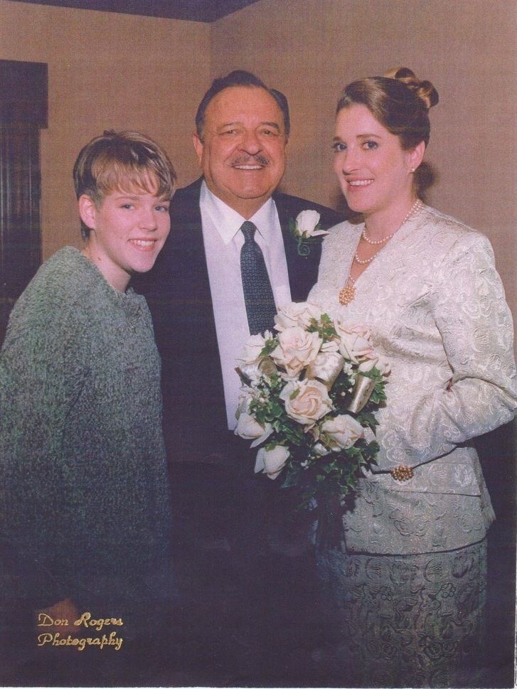 Celeste smiling and holding a bouquet and wearing a white dress while Steven Beard wearing a black coat, white long sleeves, and necktie