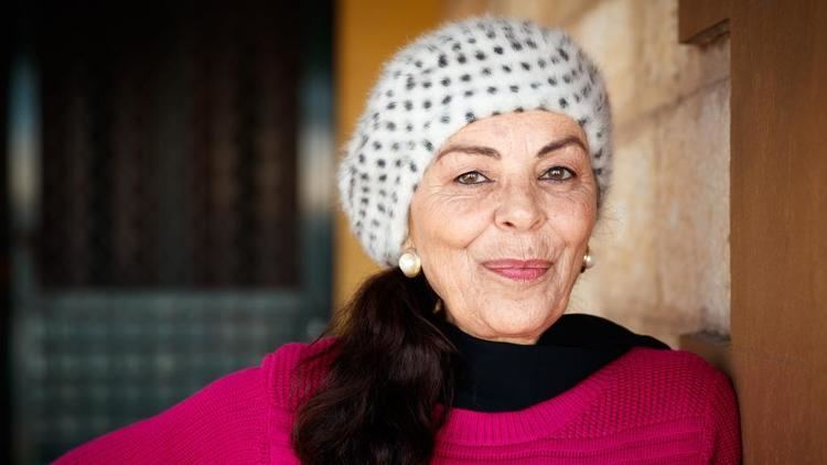 Chantal Contouri smiling with her black hair wearing a white with black dots bonnet and wearing a pearl earring, a black scarf, and a pink jacket