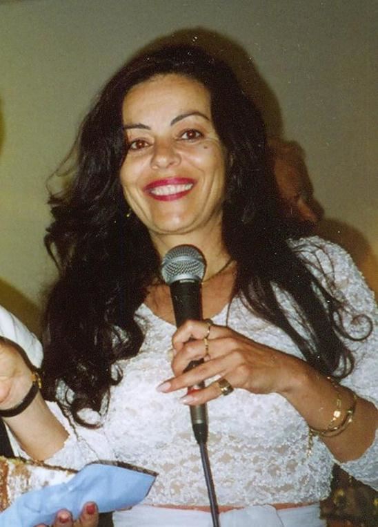 Chantal Contouri smiling while holding a microphone in her left hand, she has black wavy hair wearing an earring, necklace, bracelet on her left hand with 3 rings on its fingers, a black watch on her right wrist, and a white long sleeve dress