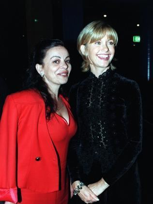 Chantal Contouri  (left) and Olivia Newton-Jon (right) are smiling together. Chantal has black hair, wearing an earring, and a visible cleavage red dress under a red coat while Olivia holds her hands together, she has short blonde hair wearing a bracelet on her right hand and a black dress