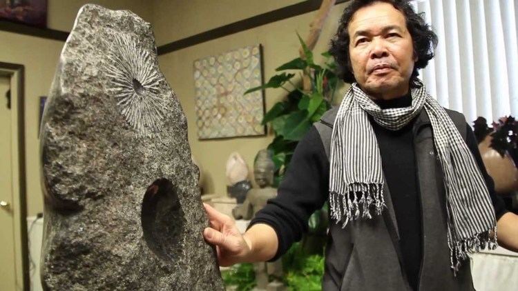 Chanthou Oeur seriously looking while touching the stone with his right hand in a stone sculpture beside him with plants and a chiral frame in the background in a clip of “Continuing the Dream” Interview Series 10 on YouTube, he has black hair, wearing a striped scarf around his neck and black long sleeve under a gray coat