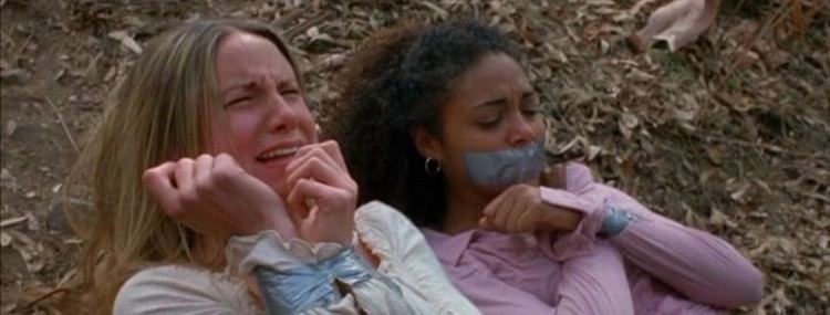 Maya Barovich as Angelica crying while both hands are tied with duct tape and lying on the ground with Chantal Degroat as Emily with hands tied together and her mouth covered with duct tape in a scene from the movie Chaos (2005 film). Maya with a blonde hair, wearing a white blouse while Chantal with curly hair, wearing a pink blouse and round silver earrings