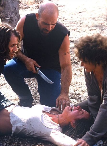 Maya Barovich as Angelica struggling and crying with her eyes closed while being held in the legs by Stephen Wozniak as Frankie, Kelly Quann as Daisy is holding both of her arms and Kevin Cage points a knife at her in a scene from the movie "Chaos" (2005 film). Maya is wearing a white blouse and a necklace, Stephen is wearing a gray jacket while Kevin with a beard, mustache and a tattoo on his right arm is wearing a black shirt and jeans
