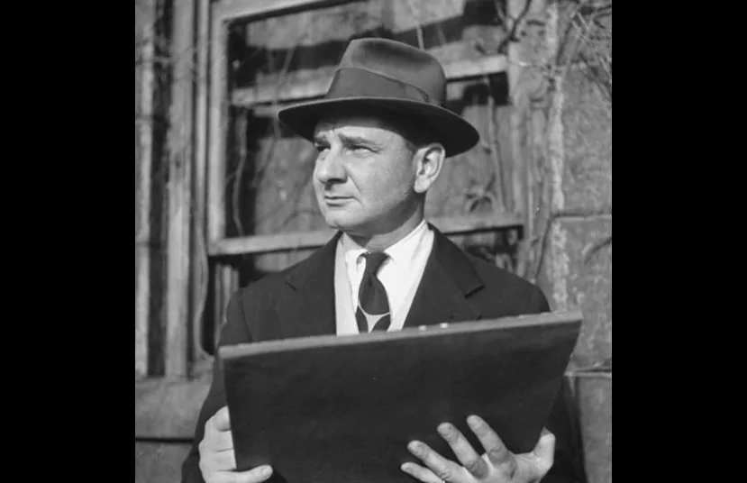 Charles Addams looking afar while wearing a hat, coat, long sleeves and necktie
