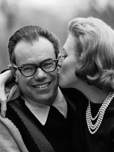 Charles Bluhdorn (left) is smiling, has black and white hair, wearing black eyeglasses, and a black and white crew neck sweater. Yvette Bluhdorn (right) is kissing her husband, has white hair, her right hand on her husband’s shoulder, wearing a pearl necklace and a black top.