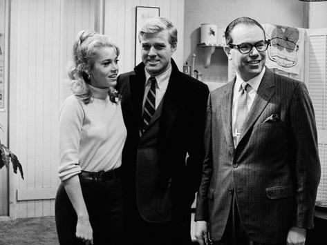 In the making of Barefoot in the Park in a room with furniture, Jane Fonda (left) smiling, has white hair, wearing a white turtle neck, black belt, and pants. Robert Redford (middle) is smiling, has white hair, left hand in his pocket, wearing a white long sleeves, black and white necktie, under a black suit and pants. Charles Bluhdorn (right) is smiling and has black hair, wearing black eyeglasses, white long sleeves, and a gray necktie under a light gray suit with lines with a handkerchief on his left pocket.