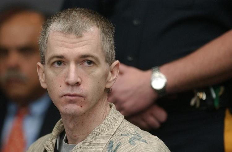 In a courtroom with a man and a police at the back, from left a man is serious sitting, has bald hair wearing a blue polo with orange necktie under a black coat, at the right is a man standing with his left hand holding his right arm, wearing a black police uniform, a silver watch and keys on his belt, in front, Charles Cullen is serious, sitting looking to his left, has brown-white hair, wearing a gray shirt under a cream-brown printed polo.