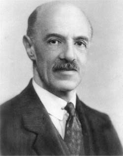 Charles Spearman with a tight-lipped smile, mustache, and bald head while wearing a long sleeve under a necktie and coat