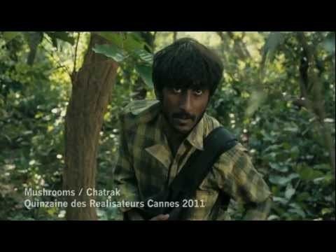 The movie scene of the Chatrak(2011) in a forest with large trees, vines and bushes, Sumeet Thakur is serious, walking, has black hair, mustache and a beard, wearing a checkered long sleeve and a black strap bag on his left shoulder. at the bottom left is a word " Mushroom / Chatrak" "Quinzaine Des Réalisateurs Cannes 2011"