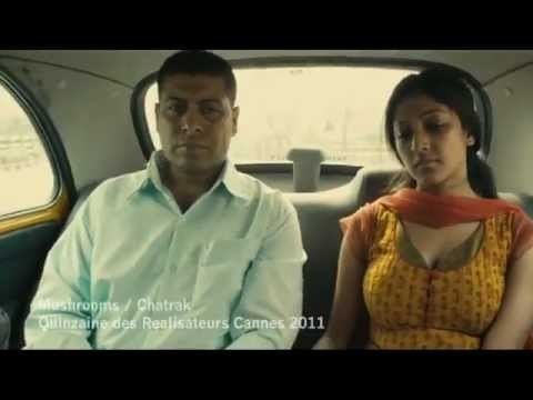 The movie scene of the Chatrak(2011) in a car with a white interior and gray leather seats, from left, Sudeep Mukherjee is serious, sitting, has black hair, wearing a white long sleeve polo, at the right, Paoli Dam is sad, looking down, eyes closed, sitting at the passenger seat, has black hair wearing a yellow cleavage showing dress, and orange scarf on her neck. at the bottom left is a word " Mushroom / Chatrak" "Quinzaine Des Réalisateurs Cannes 2011"