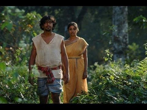 The movie scene of the Chatrak(2011) in a forest with large trees and bushes, at the left, Sumeet Thakur is serious, standing, hands down has black hair mustache and beard, wearing a ragged white top with a cloth wrapped on his waist, and a blue striped cloth at the bottom, at the right, Paoli Dam is serious, standing, has black hair, wearing a yellow saree.
