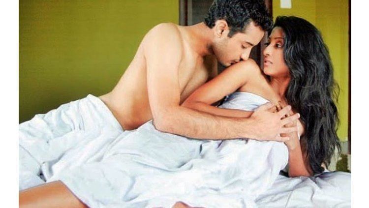 The movie scene of Hate Story (2012) in a room with yellow walls and white bed, from left is Gulshan Devaiah is leaning down with his left arm while holding the hands of Paoli, has black hair naked while under the white sheet, at the left, Paoli Dam is laying on the bed with both her hands holding the bed sheet covering her body, looking at Gulshan, has long black hair, naked.