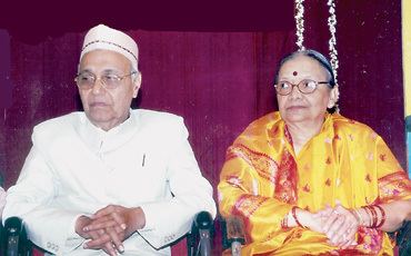 Chennaveera and Shantadevi Kanavi with serious faces while sitting on a chair. Chennaveera wearing an off-white taqiyah, eyeglasses, and a white thobe while his wife wearing eyeglasses and a yellow and red dress.