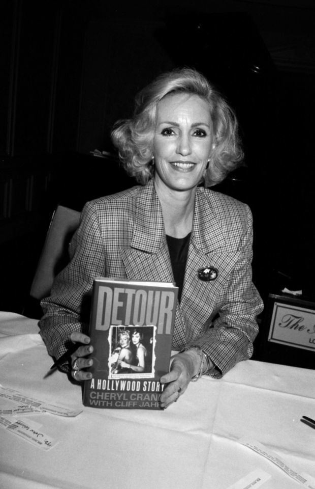 Cheryl Crane smiling and holding a book while wearing a checkered blazer and black inner top