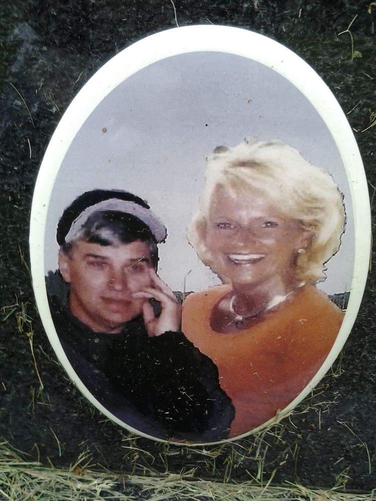 Gary Lynn (left) is serious and has black hair, his left hand on his face, wearing a black and white cap, and a black jacket. Cheryl Ann (right) is smiling, has white hair, wearing earrings on her left ear and silver necklace, and an orange top.