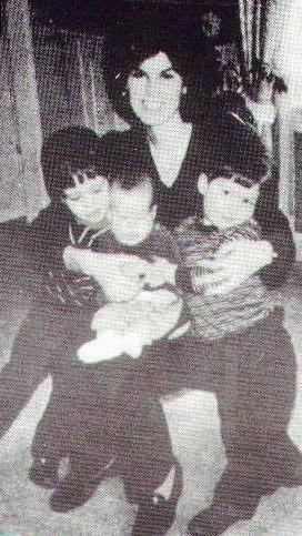 Cheryl Keeton is smiling, has black hair, holding her 3 sons, she is wearing a black dress, a boy on (left) is serious, has black hair, wearing a black long sleeve shirt, pants, and shoes, in the middle is a boy being serious, has black hair, wearing a black top, white pants, and socks, a boy on (right) is serious has black hair, wearing a black and white long sleeve shirt, black pants and shoes.