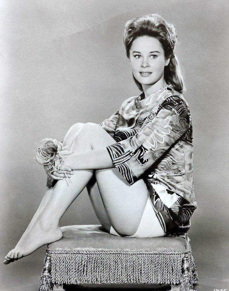 Cheryl Miller smiling while sitting on an ottoman chair and holding a flower, with shoulder-length hair, wearing a floral blouse, and white shorts.