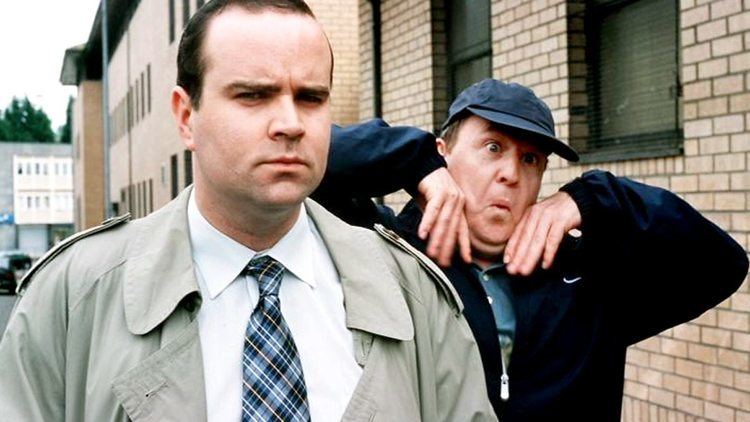 Greg Hemphill looking serious, wearing a white polo shirt and a blue checkered necktie under a gray coat together with Ford Kiernan with a wacky face, both hands on his cheeks, with wall bricks on the street in their background, wearing a blue shirt under a blue jacket, and a blue hat