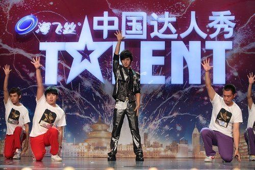 Five men in China's Got Talent. They are raising their hands with the logo of china's got Talent in the background. The two men from left to right are wearing red pants, sneakers, and white printed t-shirts. While the other two men on the right to left are wearing violet pants, sneakers, and white printed t-shirts. The man at the center is wearing a black coat, black pants, and black shoes.