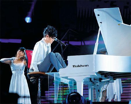 Liu Wei, China’s Got Talent winner, playing the piano with his foot and the woman at the back is playing the violin. Liu is wearing a white long sleeve, gray pants, and eyeglasses and the woman at the back is wearing a white dress