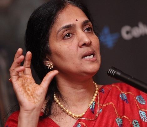 Chitra Ramkrishna is talking in front of a microphone while raising her hand, wearing earrings, a necklace, and a red traditional Indian dress called Saree.