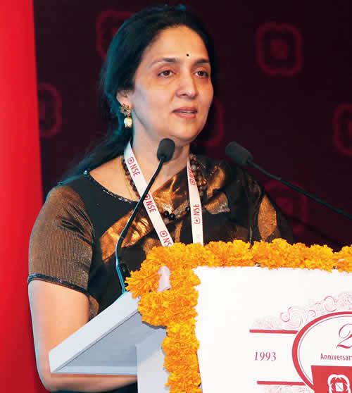 Chitra Ramkrishna with a serious face while standing beside a podium with a microphone, wearing earrings, a necklace, an ID sling, and a black and orange Indian dress called Saree.