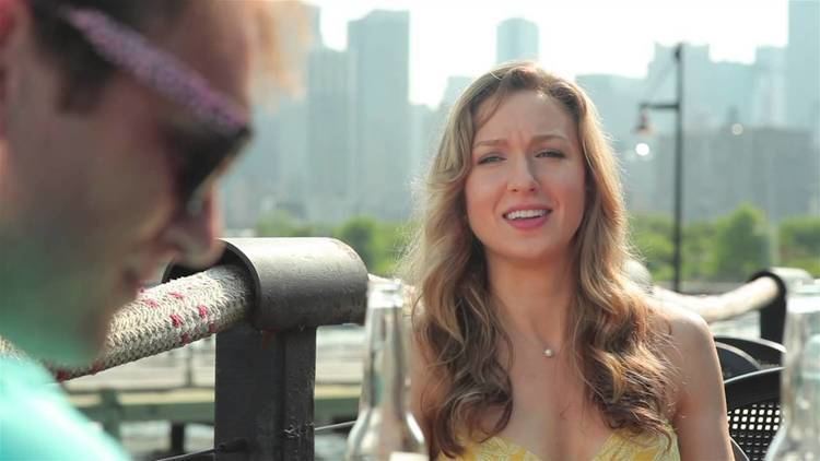 In the movie scene of THE ROTATION 2014 On the left is Chris Bayon, sitting, looking down on a  chair, has blonde brown hair wearing a pink sunglasses, and cyan shirt, At the right, Chloë Tuttle is smiling, sitting in a chair behind the table with two empty bottles, has long wavy brown hair, gray eyes, wearing a silver necklace and yellow noodle strap top.