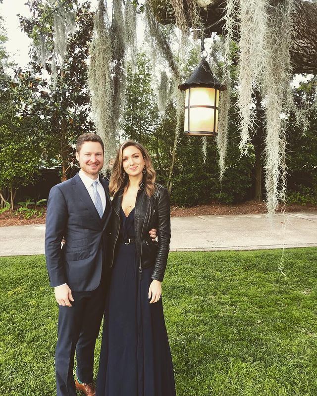 On the left, a man is smiling standing next to Chloë Tuttle with his left hand on her waist, at the back is trees, road, and a black lamp with white long shrubs, has brown hair mustache and beard, wearing a white polo with light blue tie, and black tuxedo with brown shoes,
Chloë Tuttle is smiling, standing, with her right hand on his waist, has long brown wavy hair, wearing a hoop earrings black dress black belt under a black leather jacket.