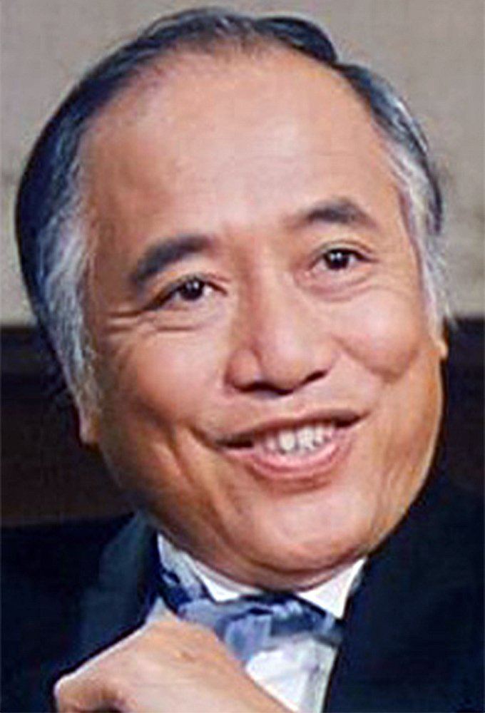 Chor Yuen smiling, wearing a black coat over white long sleeves and a blue tie.