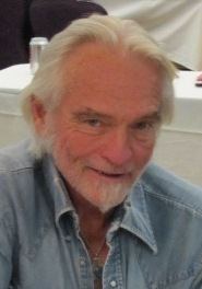 Chris Robinson is smiling, has white hair, a beard, and a mustache, wearing a silver necklace and a denim blue jacket.