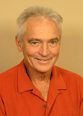 Chris Robinson is smiling, has white hair, wearing an orange shirt with buttons on chest.