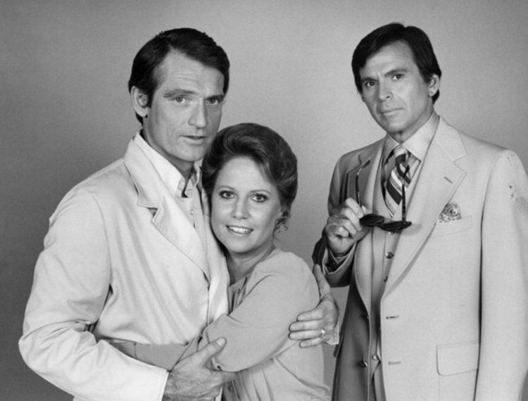 Chris Robinson (left) is serious, has black hair, and wears white long sleeves under a white suit, both hands holding Leslie. Leslie Charleson (middle) is smiling, has black hair, hugging Chris, wearing a white top. Stuart Damon (right) is serious, has black hair, right hand holding black sunglasses, wearing white long sleeves, and a necktie with lines under an off-white suit with a pocket on left.