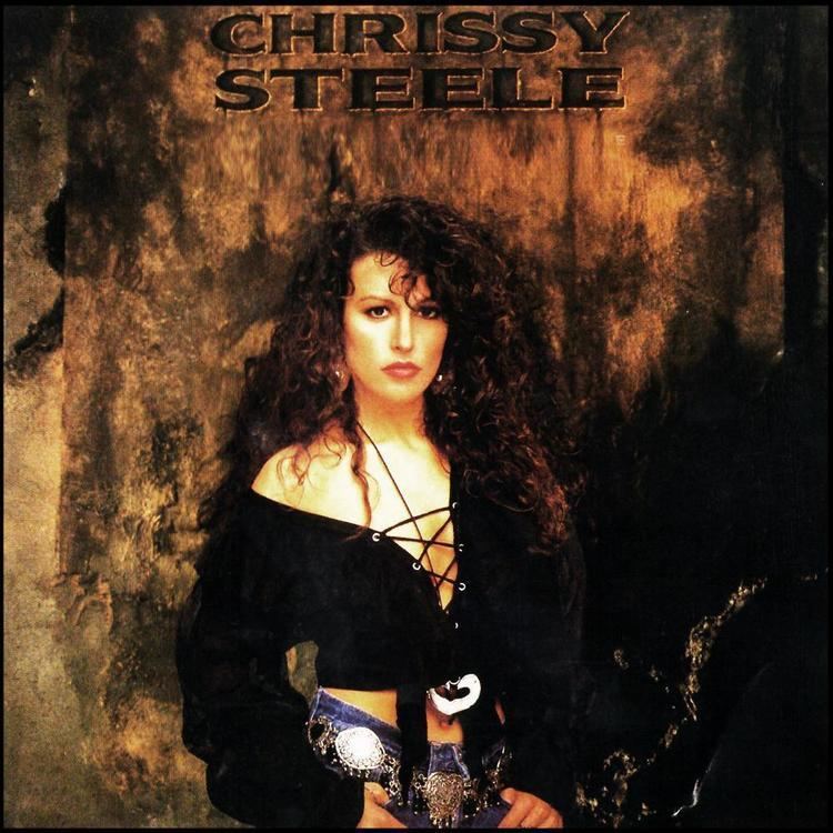 An Album cover with Chrissy Steele at the top has brown wall with a large ragged pelt, in the middle Chrissy Steele is serious standing posing with her hands thumbs on her pocket, has long brown curly hair, wearing a black long sleeve showing cleavage blouse with lace, a black belt with metal plate accessories.